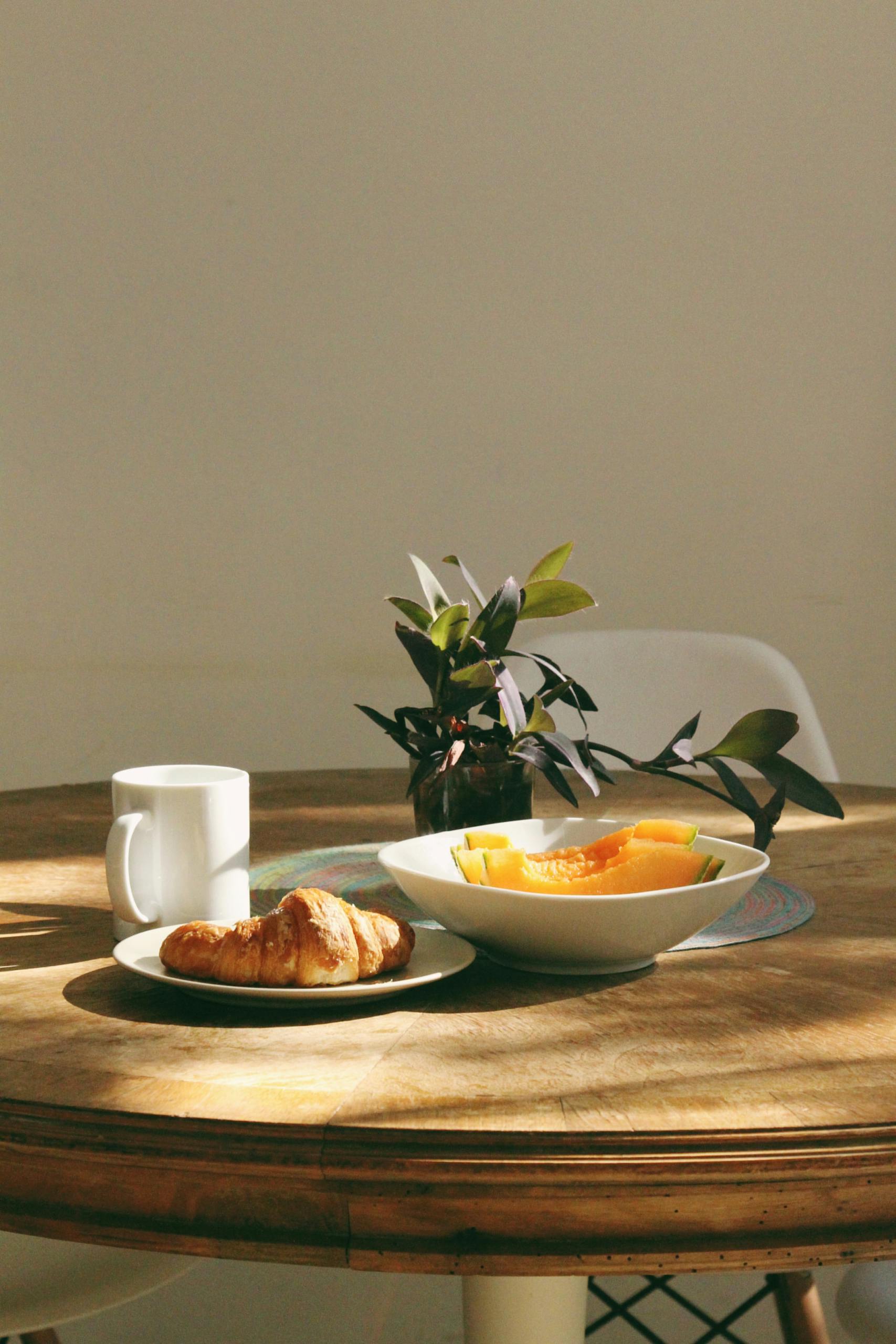 Cozy morning setup with a croissant, coffee, and fruit on a sunlit table.
